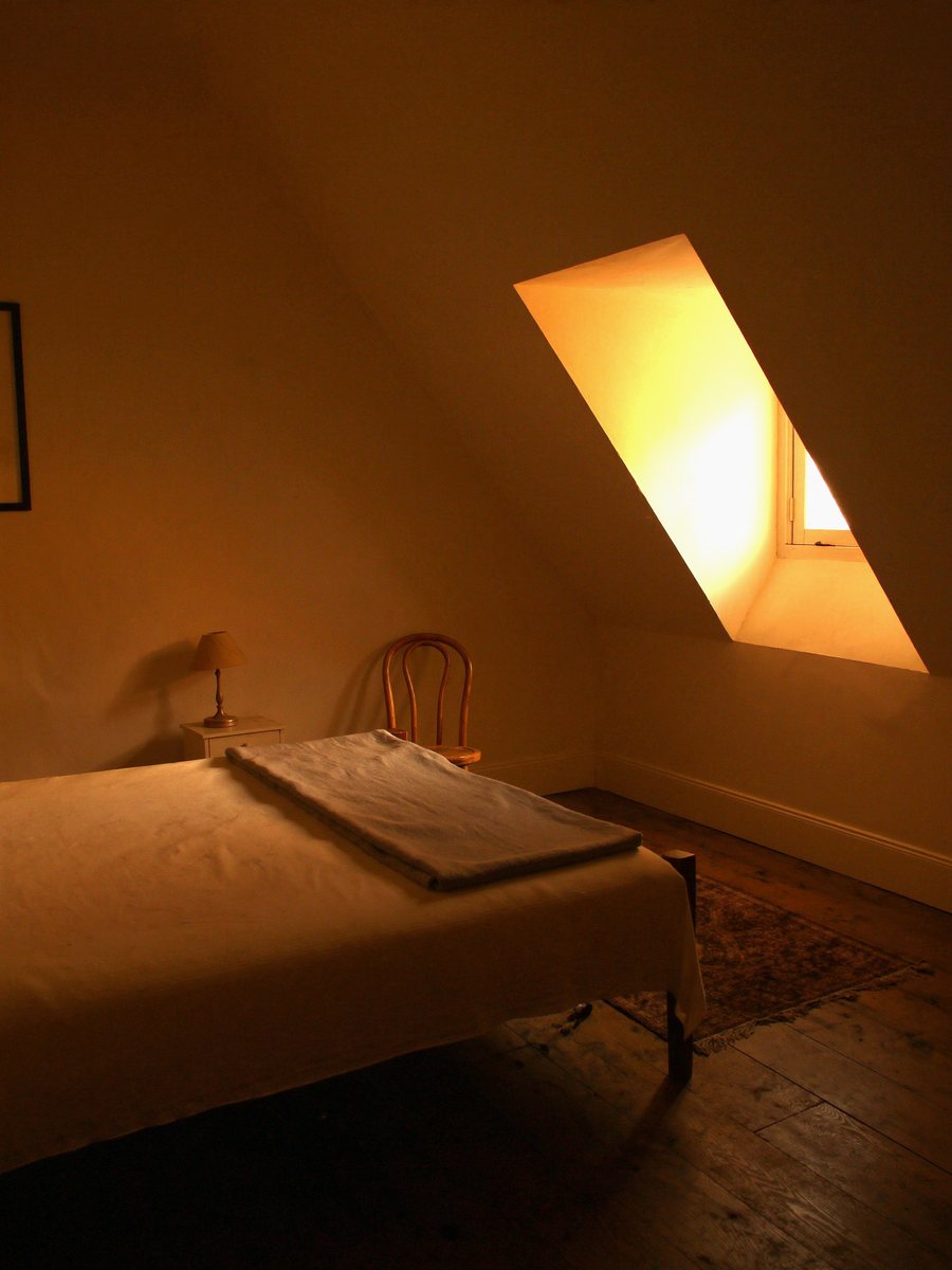 A small attic bedroom with the ceiling sloping steeply to the floor on one side, a plain bed, a bentwood chair and a dormer window letting in low evening light.