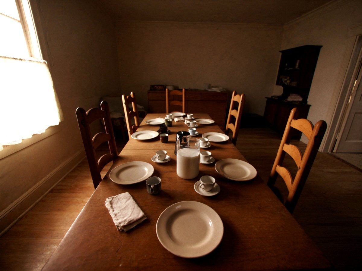 One long wooden table laid for eight with mismatched plates and cups, in a plain dining room with morning light through a lace-curtained window.