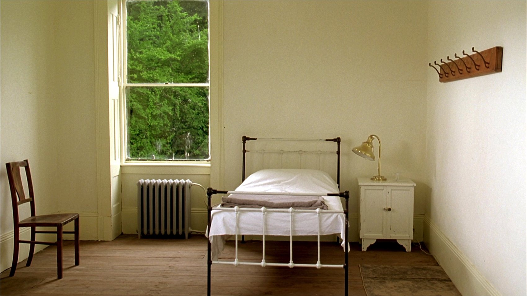 A plain guest bedroom with a white iron bedstead, a cotton coverlet, a cast iron radiator under a tall sash window, a wooden chair, a row of coat hooks and a small cupboard with a brass reading lamp on it.