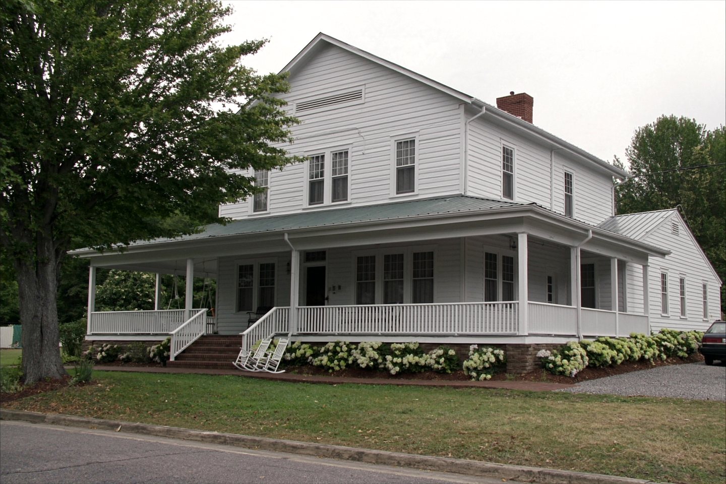A well kept two-storey white clapboard house on an overcast afternoon, with a wraparound covered porch, white rocking chairs, a green metal porch roof, hydrangea along the foundation and a gravel drive.