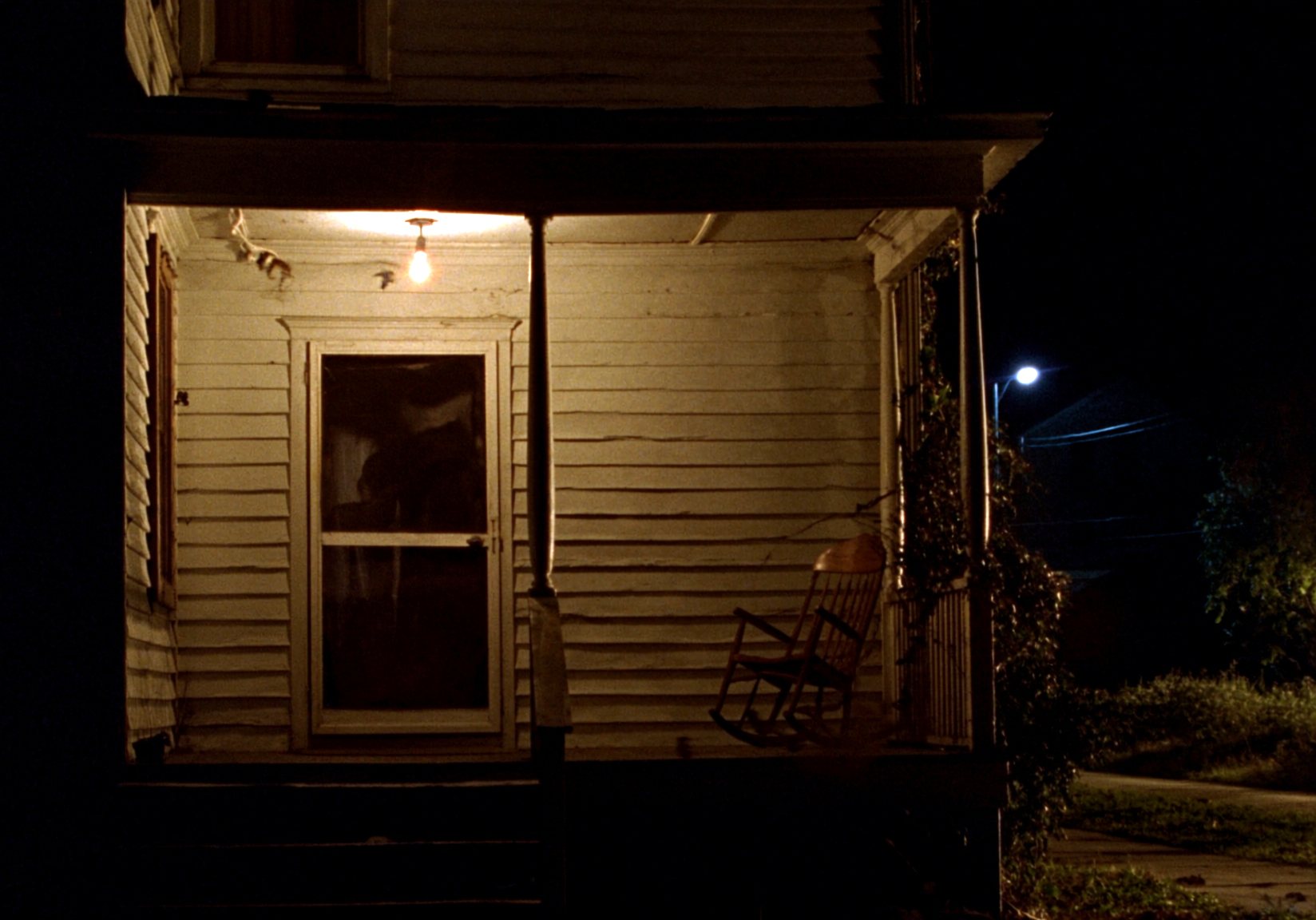 The covered front porch of an old house at night, lit by a single bare bulb over the screen door, with a wooden rocking chair and darkness beyond the porch rail.