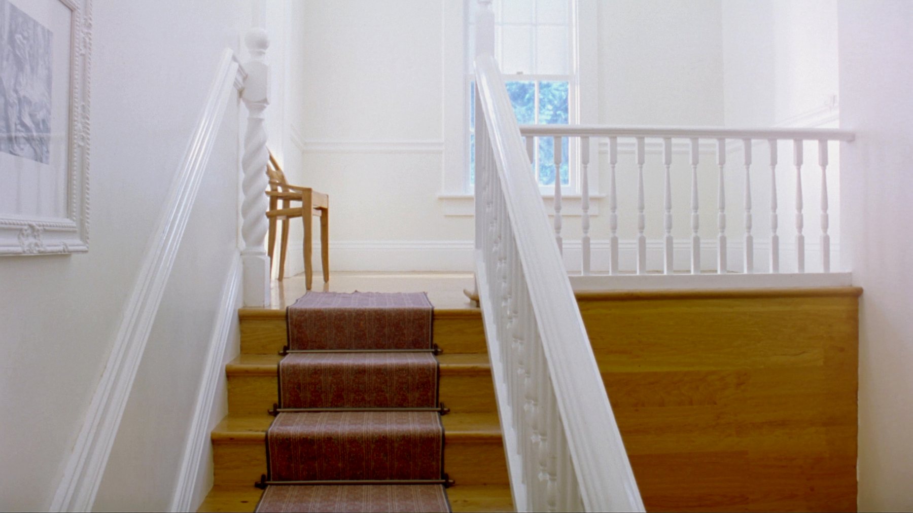 Looking up a bright wooden staircase in an old house, with a patterned stair runner, a white painted banister, and a plain wooden chair on the landing at the top.